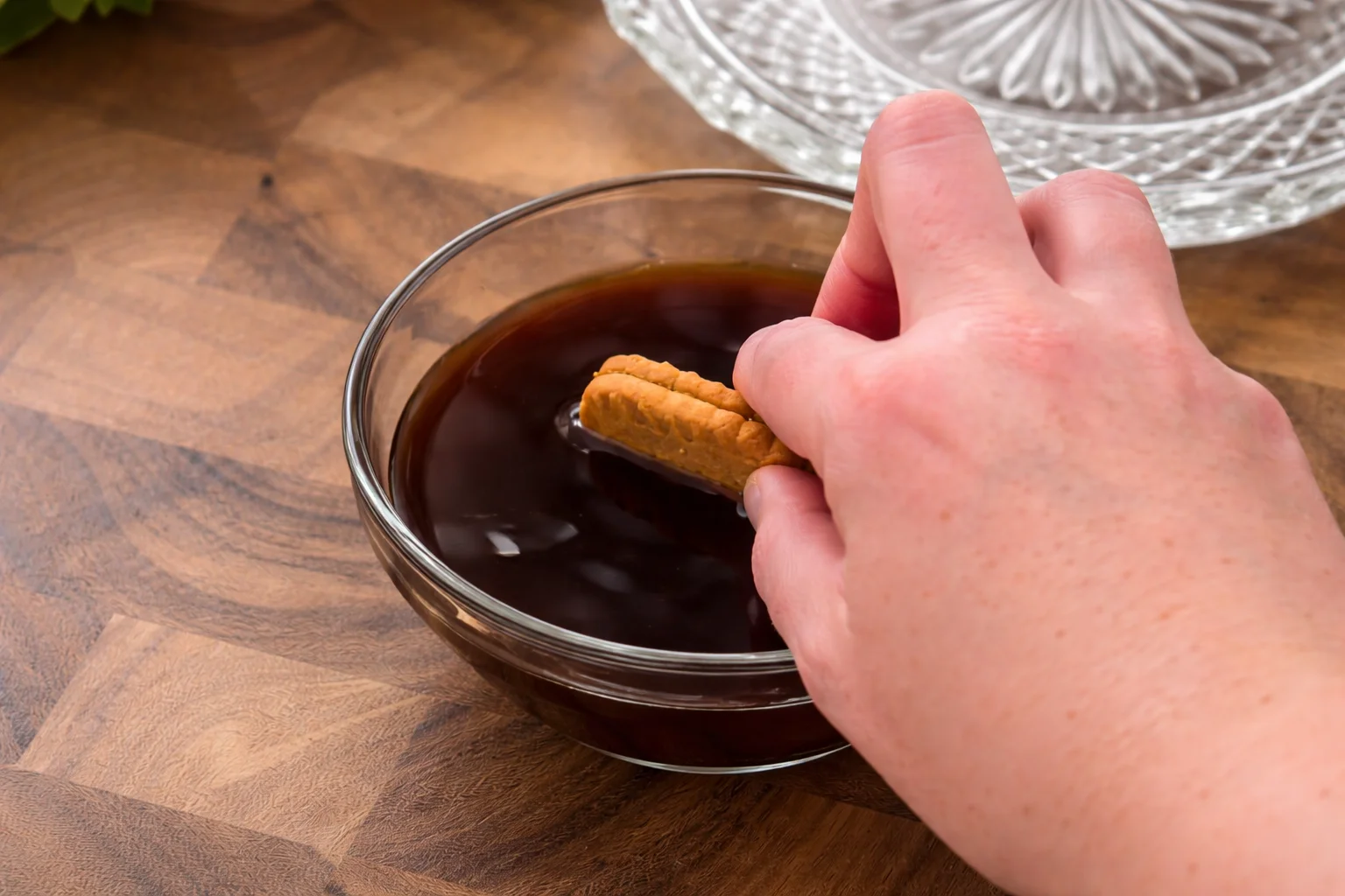 Biscoff biscuit being quickly dipped into espresso coffee in a glass bowl on a wooden kitchen surface
